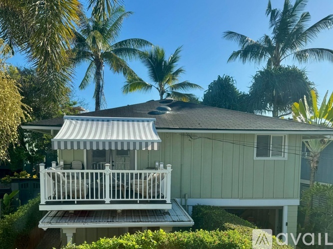 A house with a white porch and a striped awning is surrounded by greenery and palm trees.