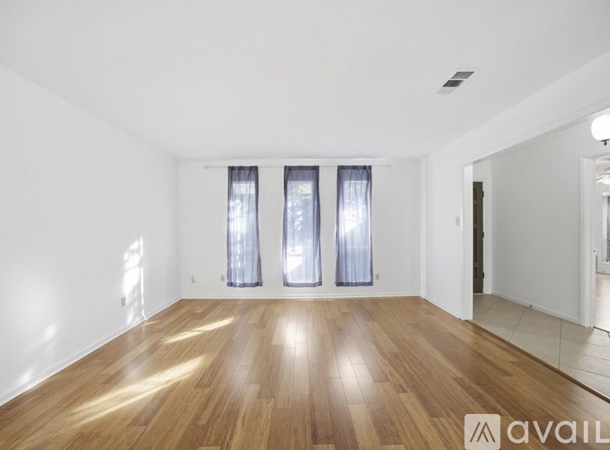 A spacious room with wooden flooring and three windows with curtains.