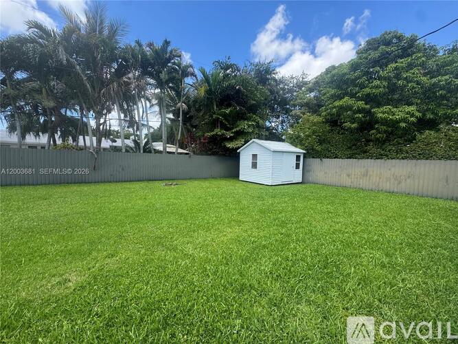 A small white house is situated in a grassy backyard with a fence and trees in the background.