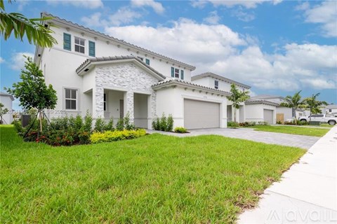 A white house with a grey garage door is surrounded by green grass and trees.