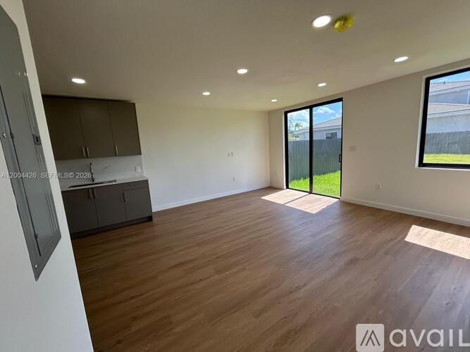 A spacious room with wooden flooring and a large window overlooking a green area.