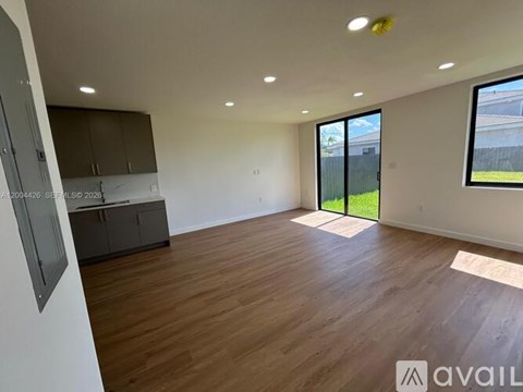 A spacious room with wooden flooring and a large window overlooking a green area.