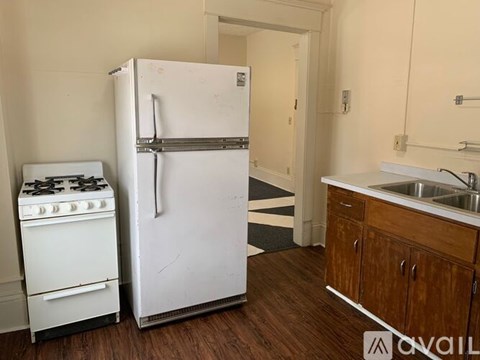 A white refrigerator stands in a kitchen with a white gas stove, wooden cabinets, and a sink.