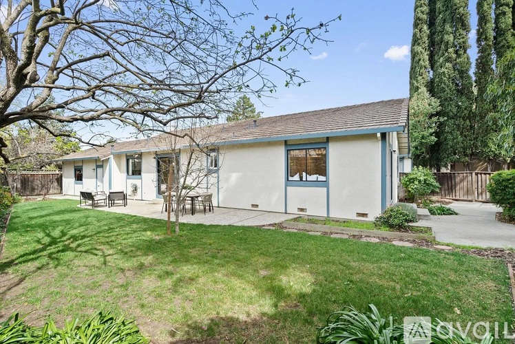 A house with a blue trim and a white roof is surrounded by a green lawn and trees.