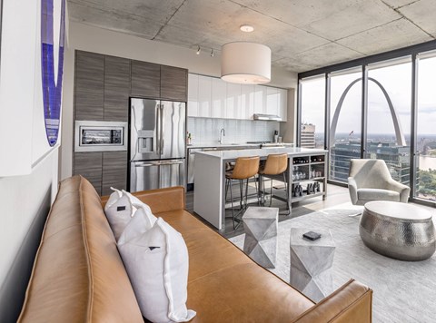A modern living room with a brown leather couch and a kitchen area with a refrigerator, sink, and stove.