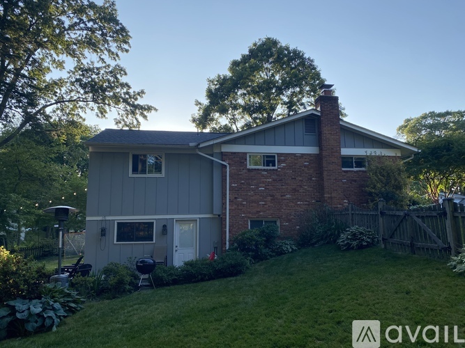 A house with a grey roof and a brick chimney is surrounded by a green lawn and trees.