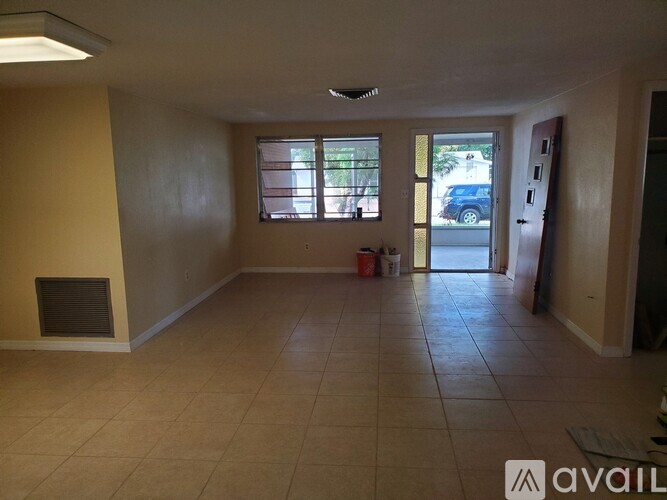 A room with beige walls and floor, a window with blinds, and a door leading to a hallway.