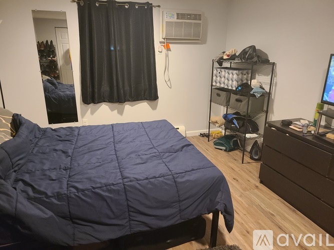 A bedroom with a bed covered in a blue comforter, a shelf with shoes, a desk with a computer, and a window with black curtains.