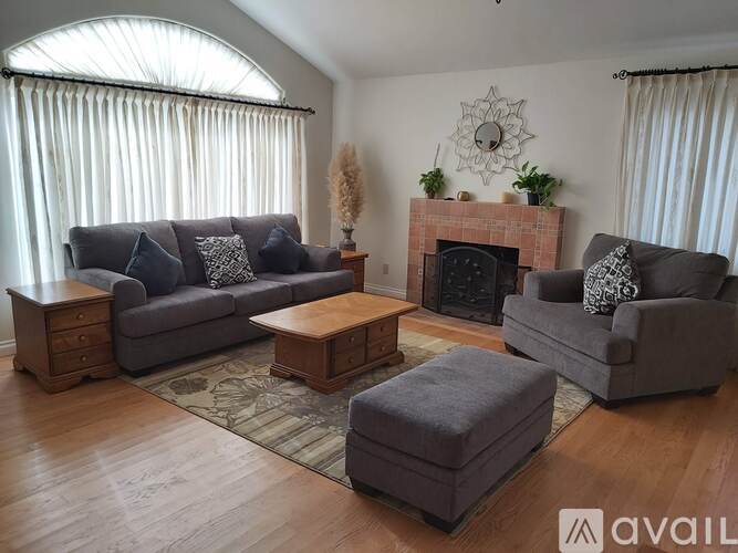 A living room with a grey couch, a grey ottoman, a wooden coffee table, a fireplace, and a large window with white curtains.