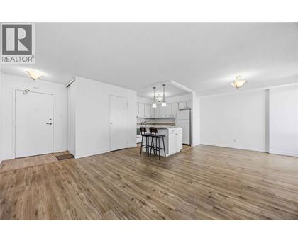 A room with wooden flooring and white walls, featuring a kitchenette with a sink and bar stools.