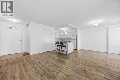 A room with wooden flooring and white walls, featuring a kitchenette with a sink and bar stools.