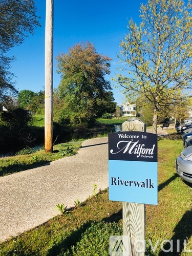 A sign welcoming people to Milford Riverwalk stands in the foreground of a grassy area.