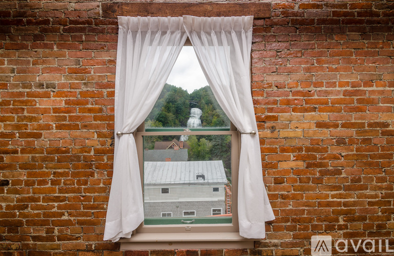 A window with white curtains and a view of a white house and trees.