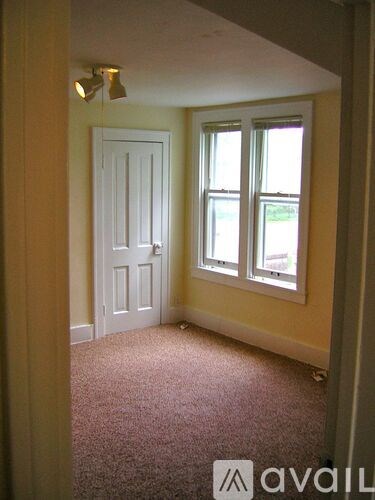A room with a carpeted floor, a white door, and a window with a view of trees outside.