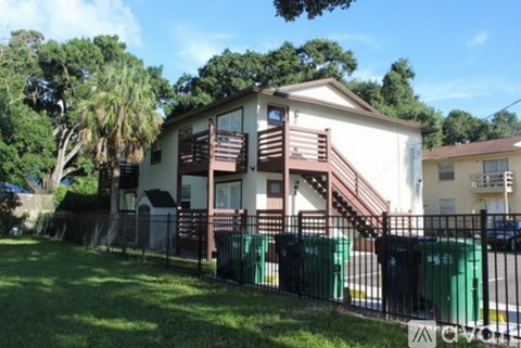 A white house with a brown deck and a black fence with green trash cans in front.