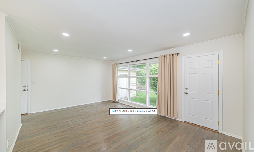 A spacious room with wooden flooring and white walls, featuring a large window with a view of greenery outside.