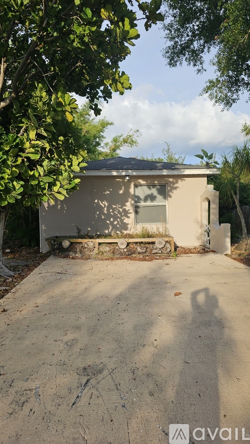A small, beige building with a dark roof is surrounded by trees and has a concrete driveway in front.