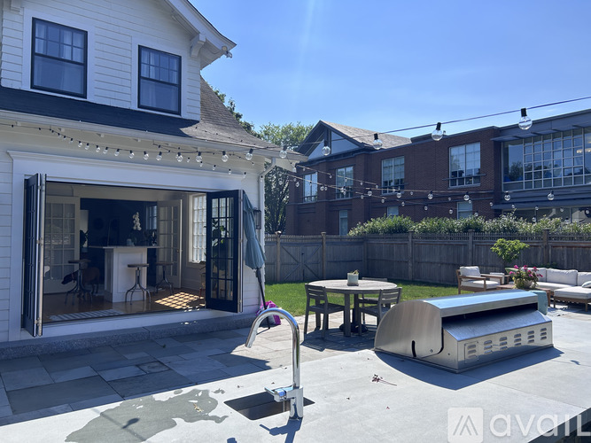 A patio with a hot tub and a grill is surrounded by a white house and a brick building.