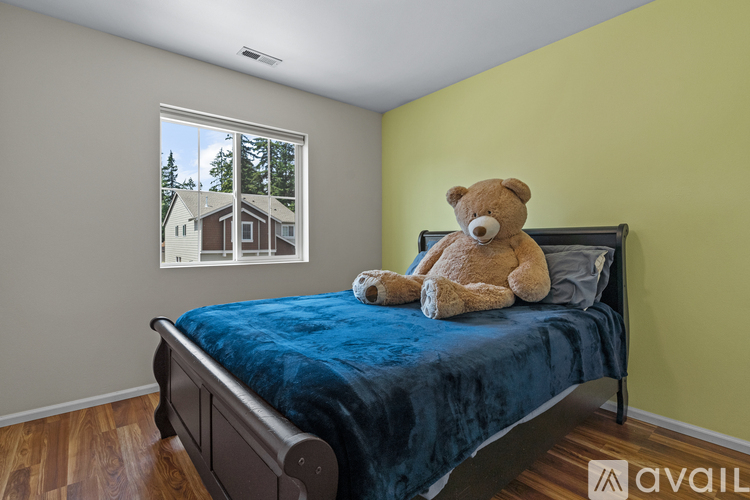 A teddy bear sits on a bed in a room with a window showing a house and trees outside.