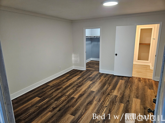 A bedroom with wooden flooring and a full bathroom visible in the background.