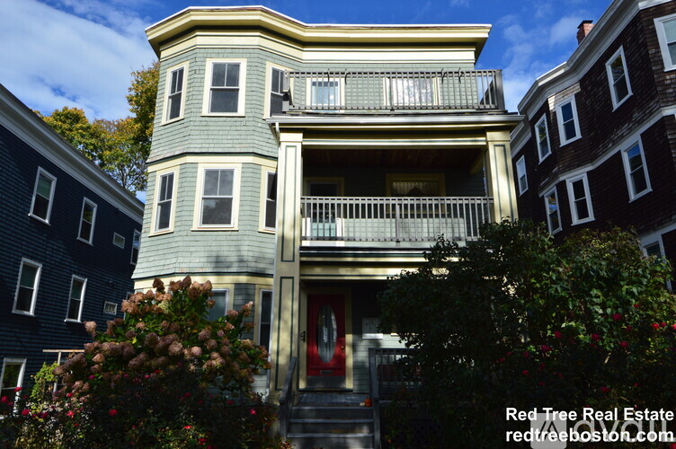 A house with a red door and a balcony is surrounded by trees and bushes.