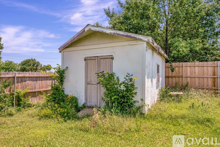 A small white shed with a brown door is surrounded by a grassy area and a wooden fence.