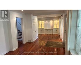 A spacious room with wooden floors and white walls, featuring a staircase and a row of windows.