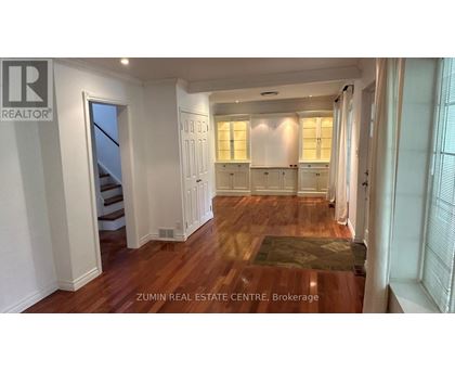 A spacious room with wooden floors and white walls, featuring a staircase and a row of windows.