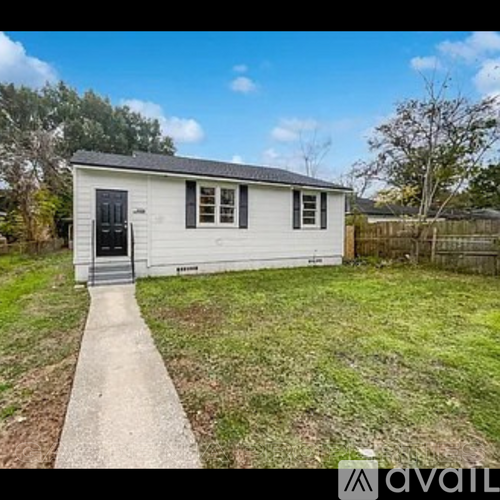 A small white house with a black door and windows is surrounded by a grassy area and a wooden fence.
