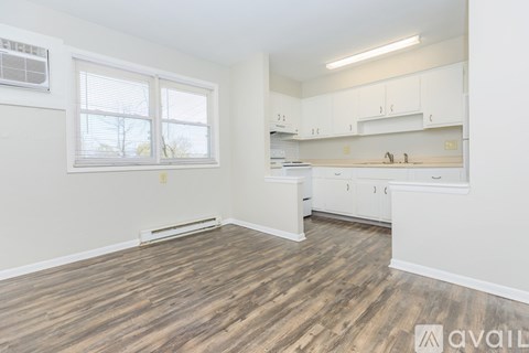 A room with wooden flooring and white walls, featuring a window with blinds, a white air conditioning unit, and a kitchenette with white cabinets.
