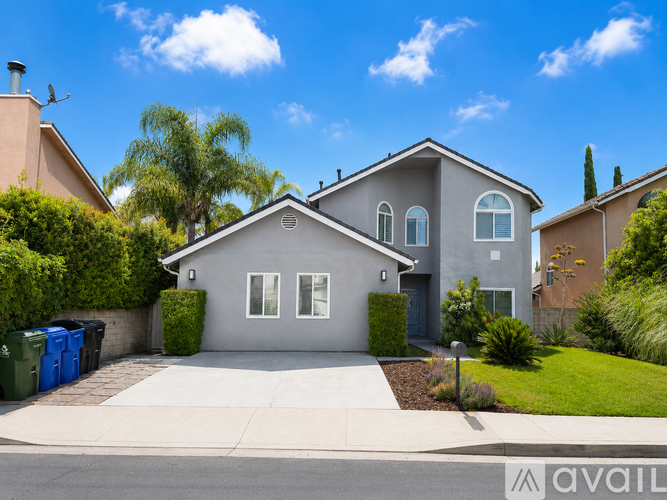 A house with a grey exterior and a white roof is surrounded by greenery and has a clear blue sky above.
