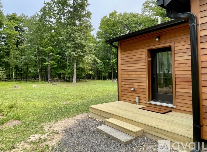 A small wooden cabin with a black roof is surrounded by a gravel path and a grassy area with trees in the background.