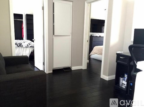A black couch sits in a living room with a white door and a bed in the background.