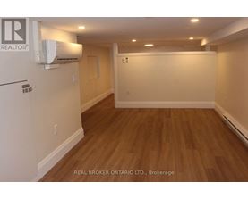 A room with wooden flooring and a white wall with a Real Broker logo.