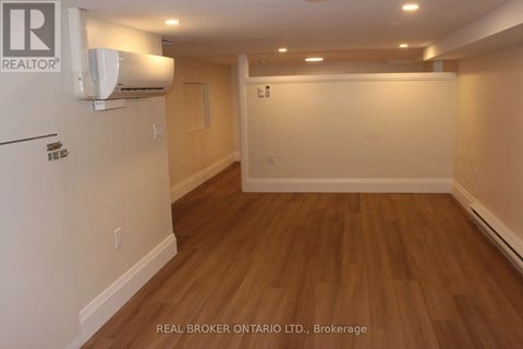 A room with wooden flooring and a white wall with a Real Broker logo.
