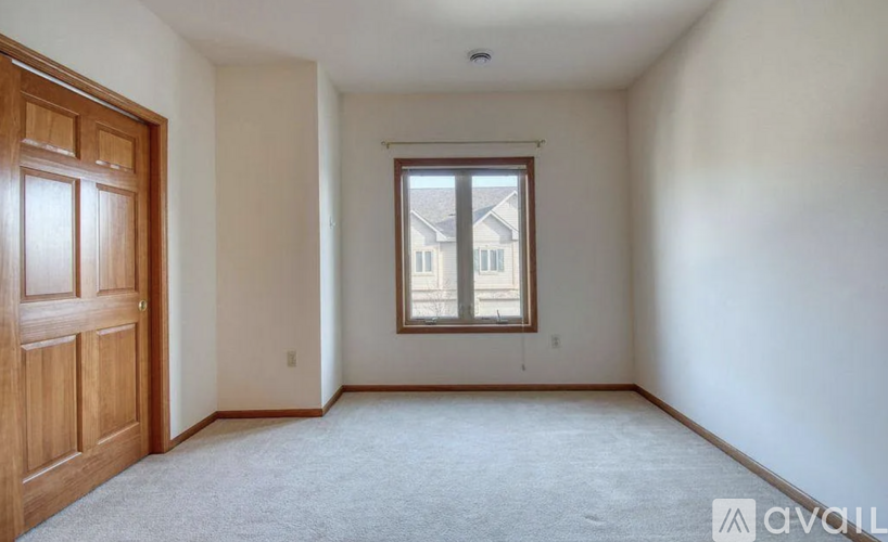 A room with a carpeted floor, a wooden door, and a window with a view of a building outside.