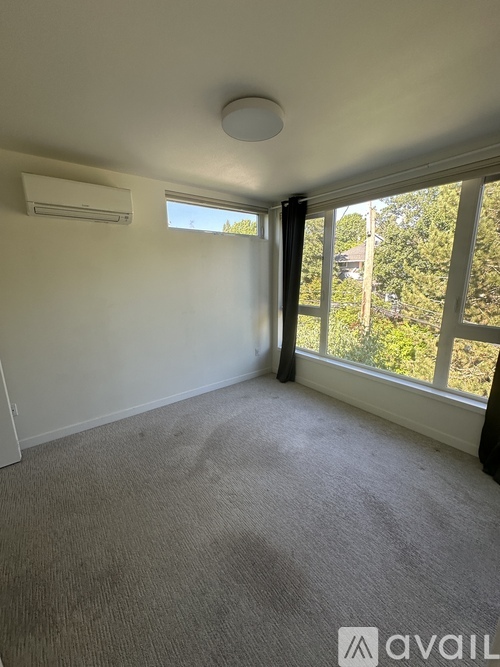 A room with a carpeted floor, a window with a view of trees, and an air conditioner on the wall.