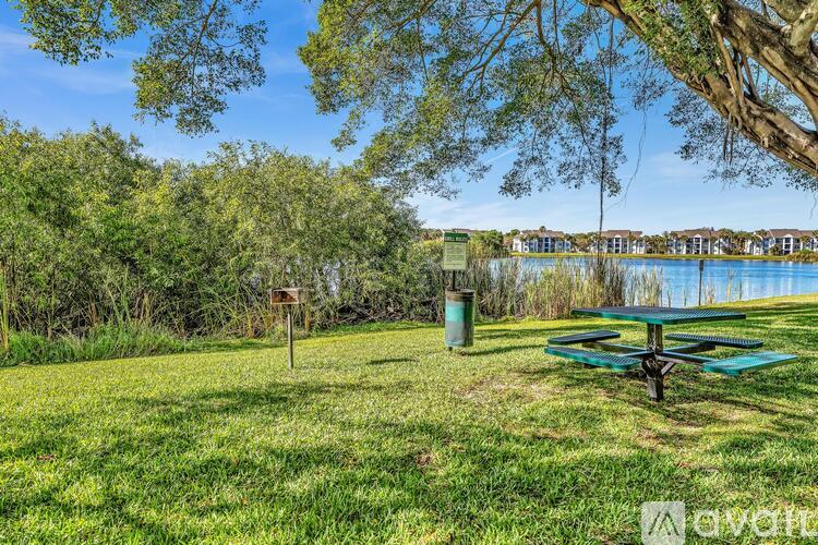 A picnic table sits in a grassy field with a lake and houses in the background.