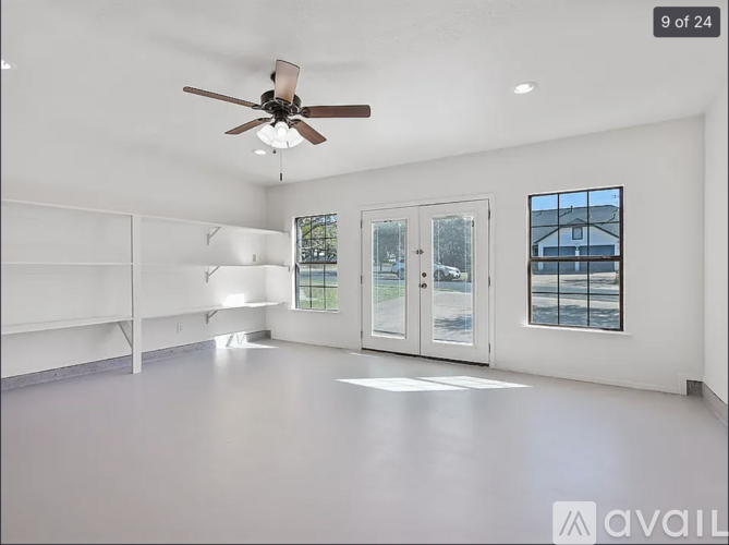 A spacious empty room with a ceiling fan and a window showing a view of a house.