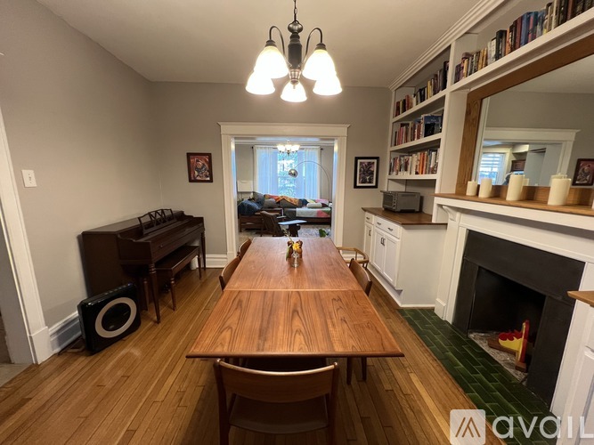 A dining room with a wooden table and chairs, a piano, a fireplace, and a bookshelf with books.