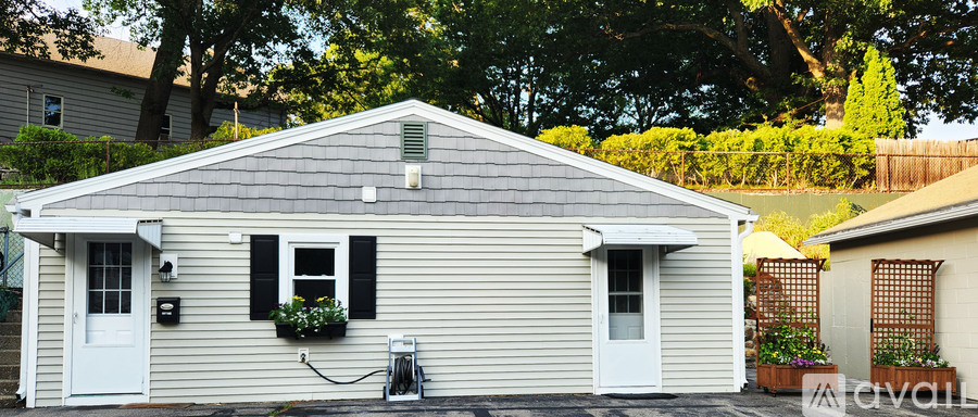 A small house with a grey roof and white walls is surrounded by greenery.