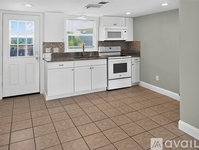 A kitchen with white cabinets and appliances, a brown tile floor, and a window with a view of trees.