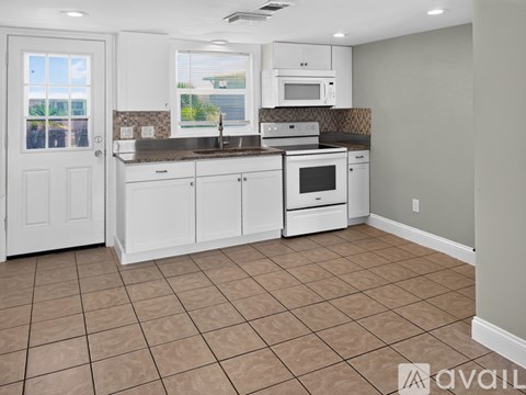 A kitchen with white cabinets and appliances, a brown tile floor, and a window with a view of trees.