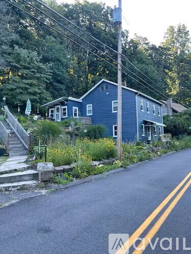 A blue house with a white door and windows is surrounded by greenery and a road in front.