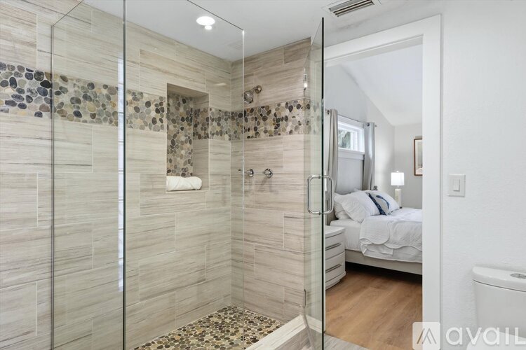 A bathroom with a glass shower stall and a bedroom with a bed and nightstand.