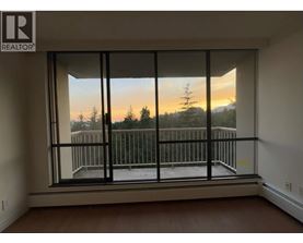 A room with a large window overlooking a balcony with a view of trees and a sunset.
