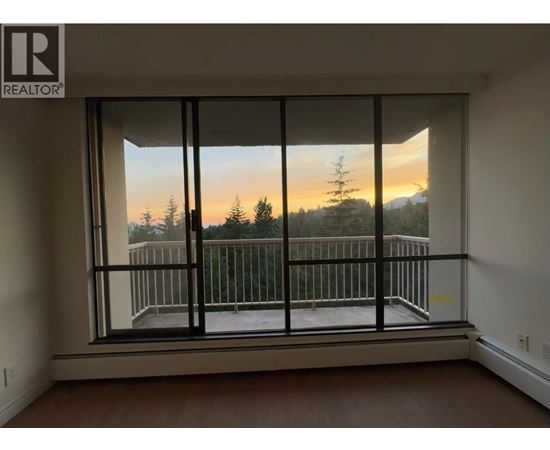 A room with a large window overlooking a balcony with a view of trees and a sunset.