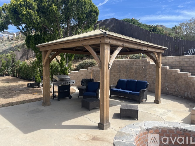 A wooden gazebo with blue cushioned seating is surrounded by a stone wall and trees.