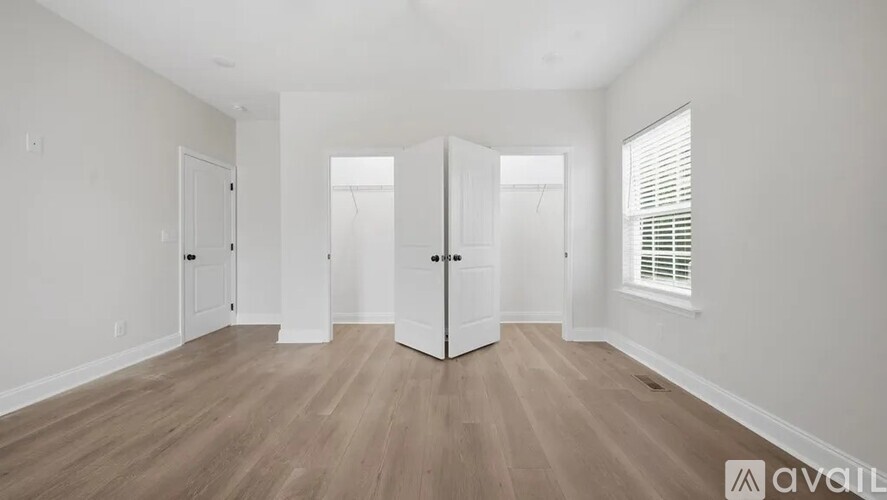 A room with wooden flooring and white walls, with a large open space and a window with blinds.