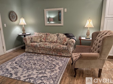 A floral sofa sits in a living room with a patterned rug and a chair with a quilted back.